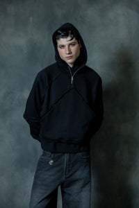 Portrait of a man wearing a premium black cotton zip-neck hoodie with the hood up, showcasing a relaxed fit and minimal urban streetwear aesthetic