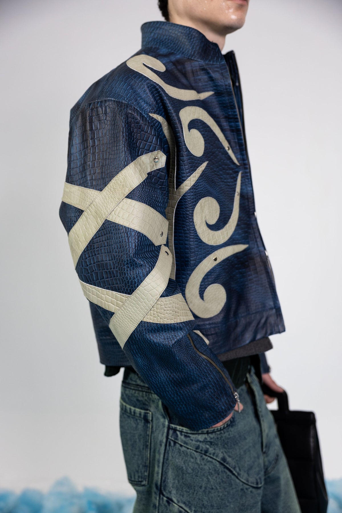 Side profile of men's blue embossed leather jacket, showcasing abstract beige appliqué design on sleeves and metal rivets