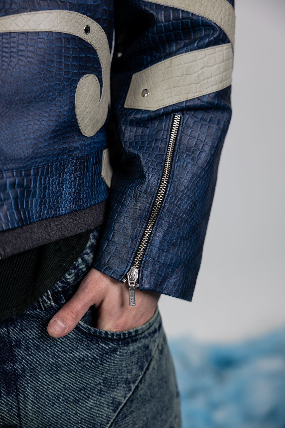 Close-up of men's blue embossed leather sleeve featuring beige appliqué patchwork and metal rivet detailing