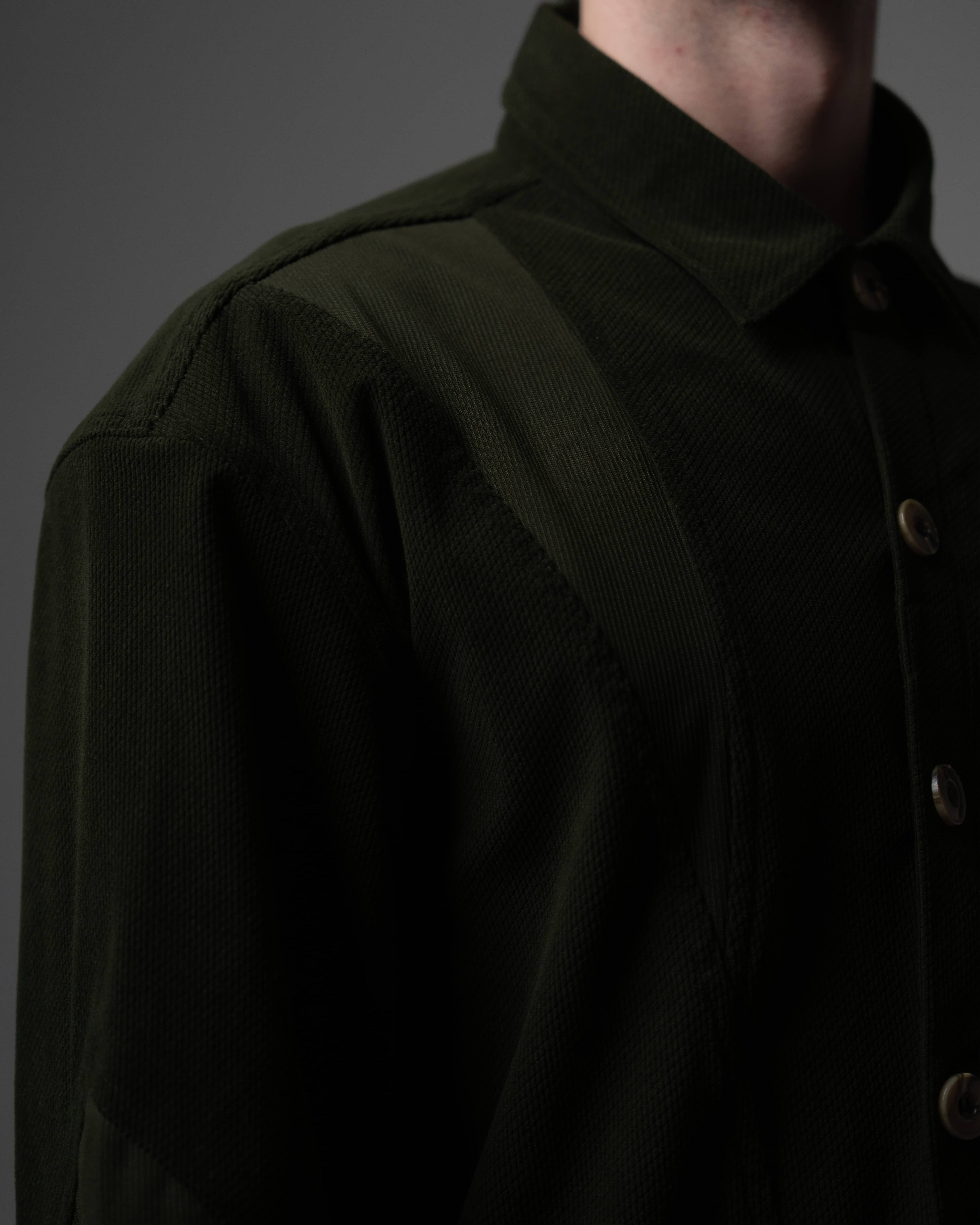 Close-up detail of the shoulder seam and stitched button on a dark olive textured shirt